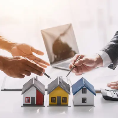 Three model houses on table with two people's hands gesturing with pens above the houses.