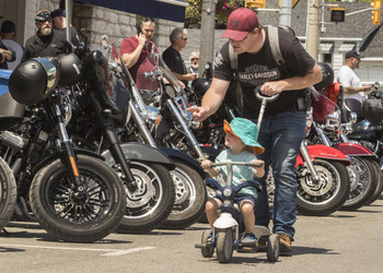 A young child sits in a stroller and is pushed by an adult male. They look at motorcycles lined up on display along the road.