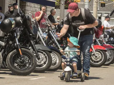 A young child sits in a stroller and is pushed by an adult male. They look at motorcycles lined up on display along the road.