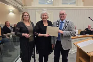 Photo of three people standing and facing the camera. The centre person is holding an envelope.